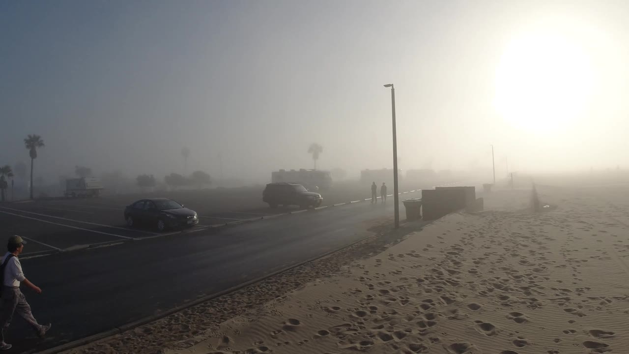 A pedestrian appears in the foreground of the Huntington Beach, California street scene, walking past sand, parked cars, palm trees, and strong sun haze.
