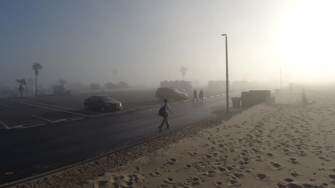 A person walks across the dark road in Huntington Beach while other distant figures stand near parked cars under fog and strong morning light.