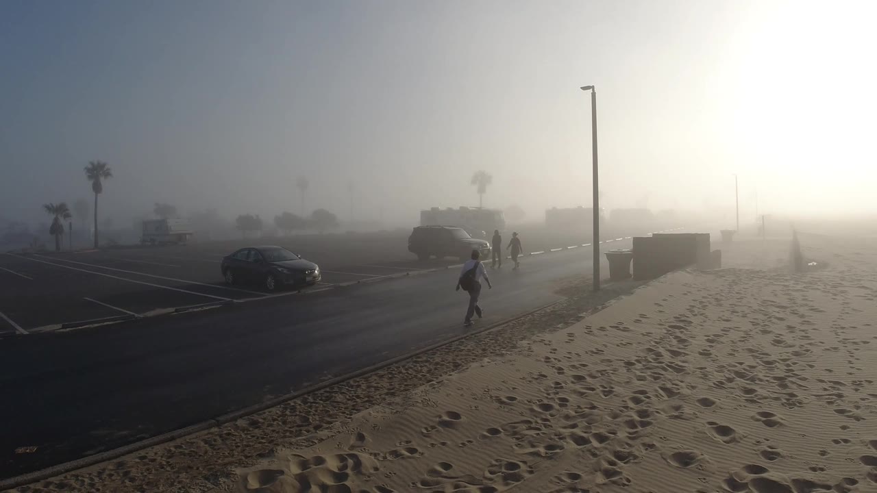 Several distant people move near a foggy Huntington Beach parking lot, with a dark car on the left, sand in the foreground, and bright sun washing the right edge.