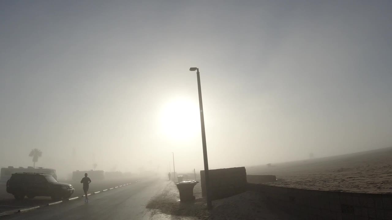 Huntington Beach street in dense fog with a parked car at left, a distant person walking, a tall streetlight, sand at right, and heavy sun glare.