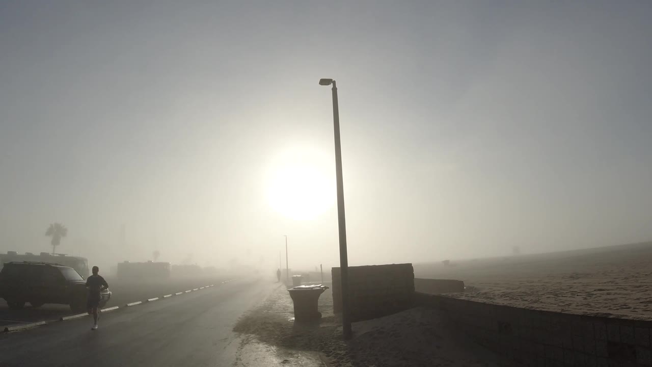 A person walks near the right side of the foggy Huntington Beach street while a parked car sits near the curb and the bright sun washes out much of the background.