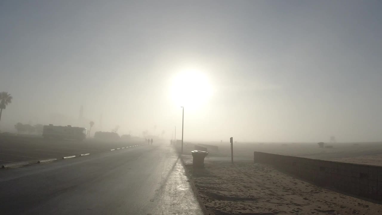 Huntington Beach beach road under blinding backlight, with dry sand at right, a low wall, distant palms, and tiny distant pedestrians softened by haze.