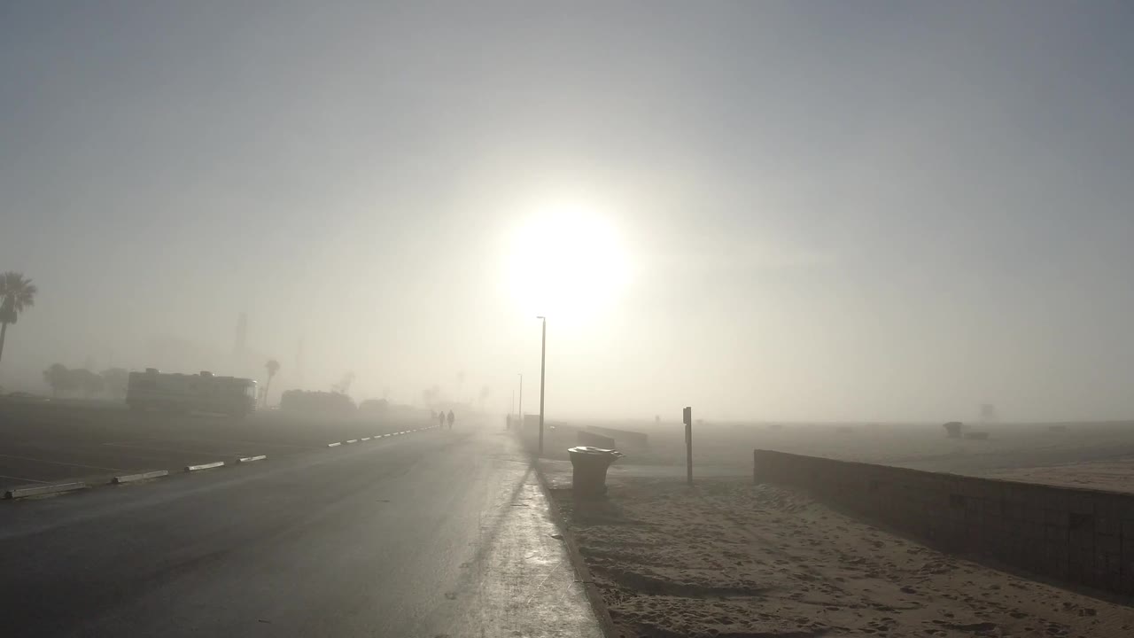 A bright Huntington Beach road scene shows the sun flaring above the pavement, with sand, a trash can, a roadside post, palm silhouettes, and distant people ahead.