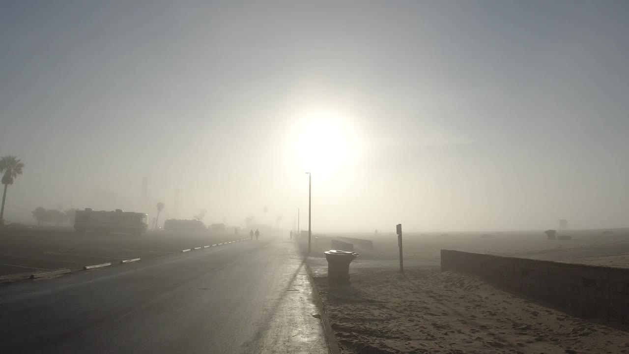 Hazy beach road with sun glare and distant pedestrians.