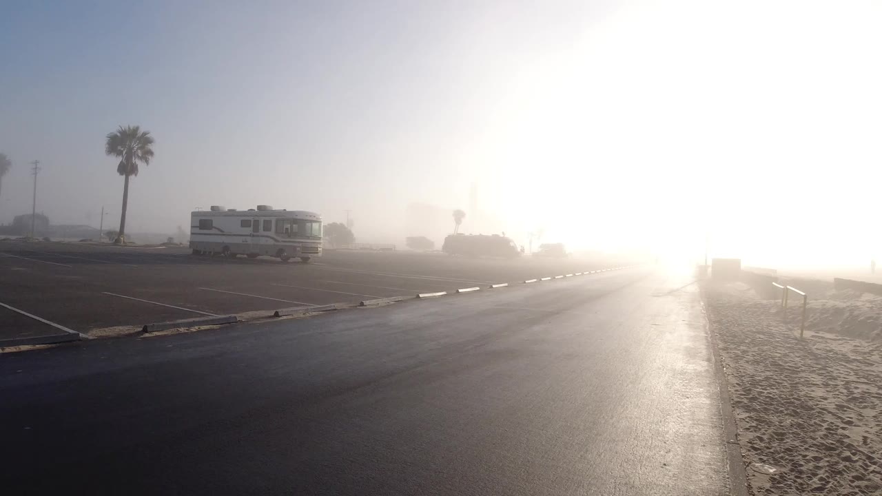 At 0:00, a hazy Huntington Beach parking area shows wet pavement leading toward bright sun, with sand at right, an RV and palm trees at left, and no visible people.