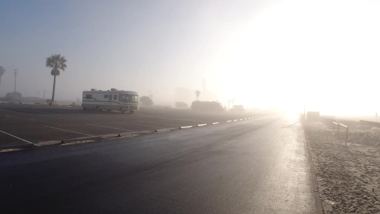 Sun glare washes over wet pavement near beach sand, with an RV and palm trees in haze.