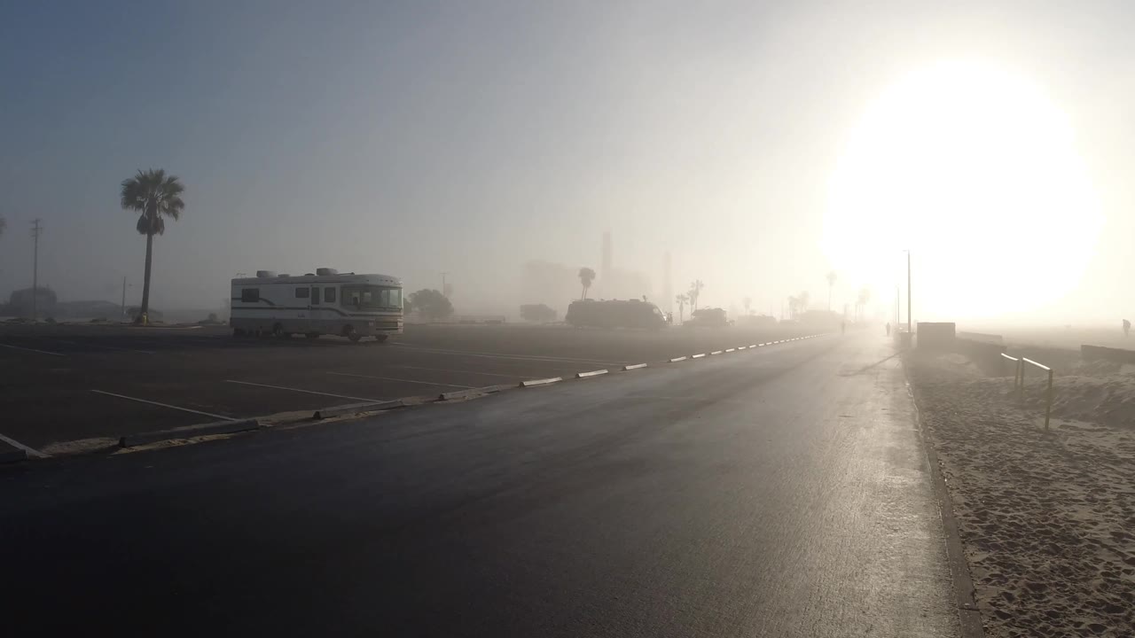 At 0:02, intense sun dominates the right side of the Huntington Beach frame while the RV, palm trees, railings, sand, and dark pavement remain visible through haze, with no visible people.
