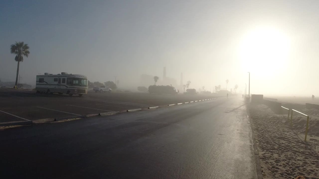 Foggy Huntington Beach road beside sand with a parked RV on the left, a palm tree, bright sun glare on the right, and no visible people.