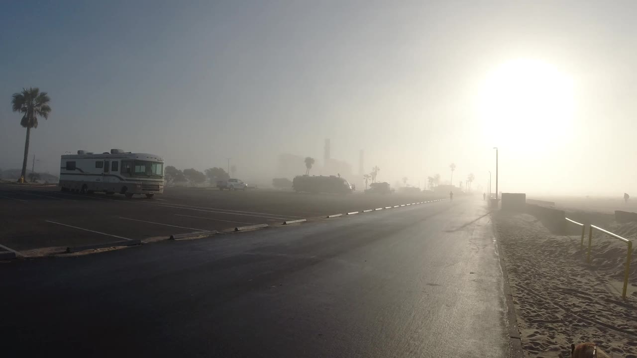 Huntington Beach fog softens the road, parked RV, sand, distant palm shapes, and bright white sun, with no visible people in the frame.