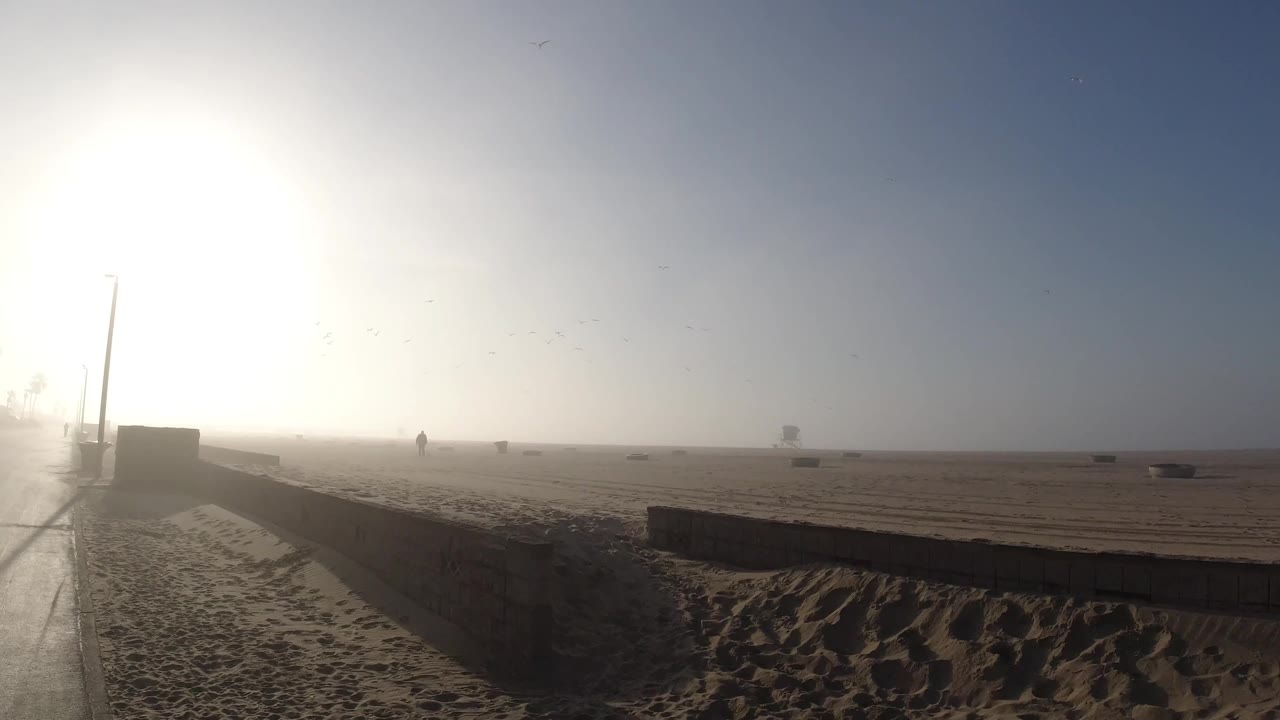 A foggy Huntington Beach morning view shows wet pavement, sand, a low beach wall, a lamp post, and bright sun glare with no clearly visible people.