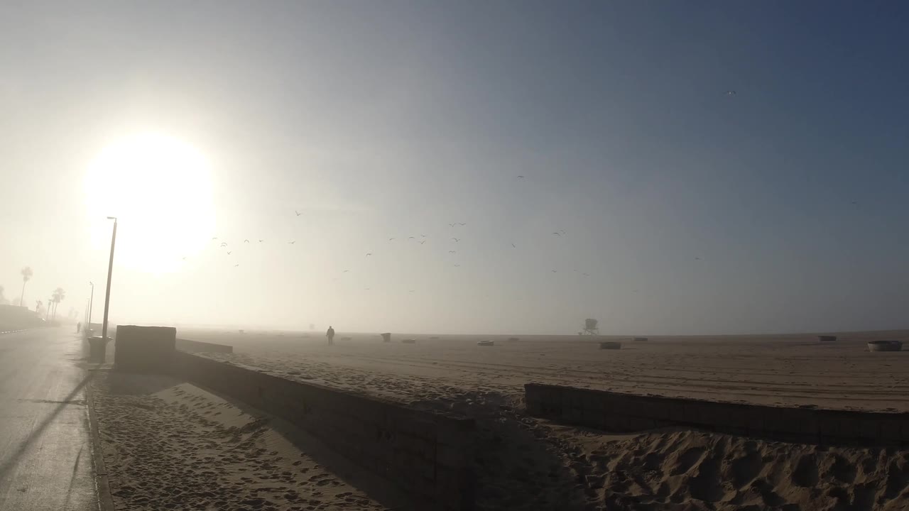 The beach remains washed in morning fog, with a lamp post and low wall silhouetted near the road and small dark objects scattered across the sand.