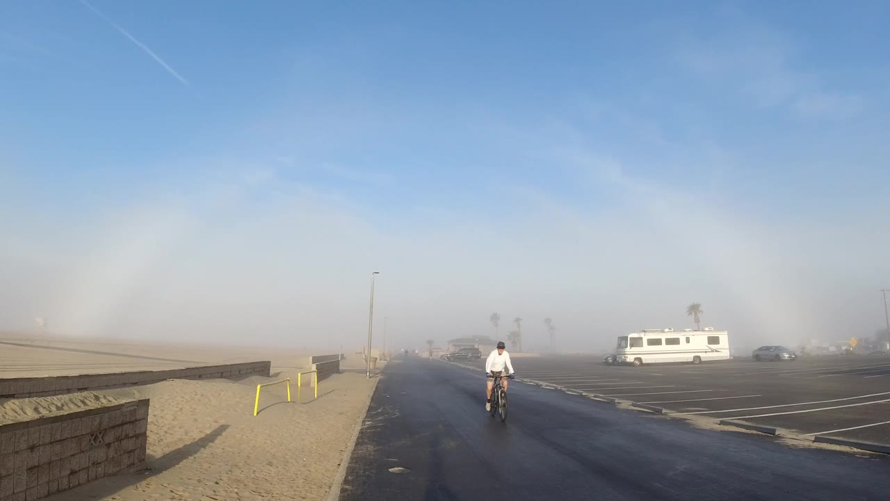 The cyclist remains near the center of the Huntington Beach roadway as the pale sky arc sits above haze, parked cars, a white RV, sandy ground, and yellow bollards.