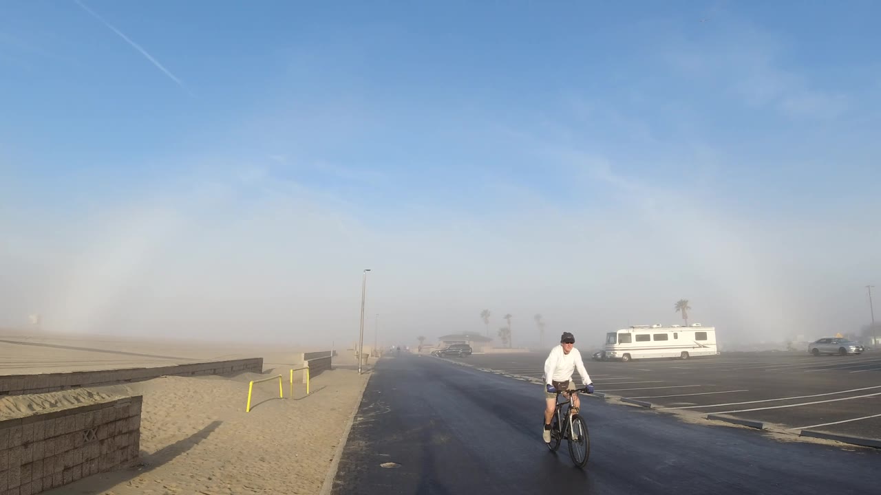 A cyclist passes near the right edge of the Huntington Beach frame while the wet-looking road, sandy shoulder, white RV, parked cars, haze, and blue sky with a faint arc remain visible.