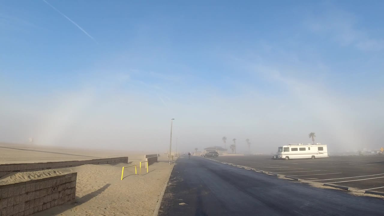 Huntington Beach roadside scene with a wet-looking road, sandy foreground, low block wall, hazy background palms, a white van in the parking area, and no visible people.