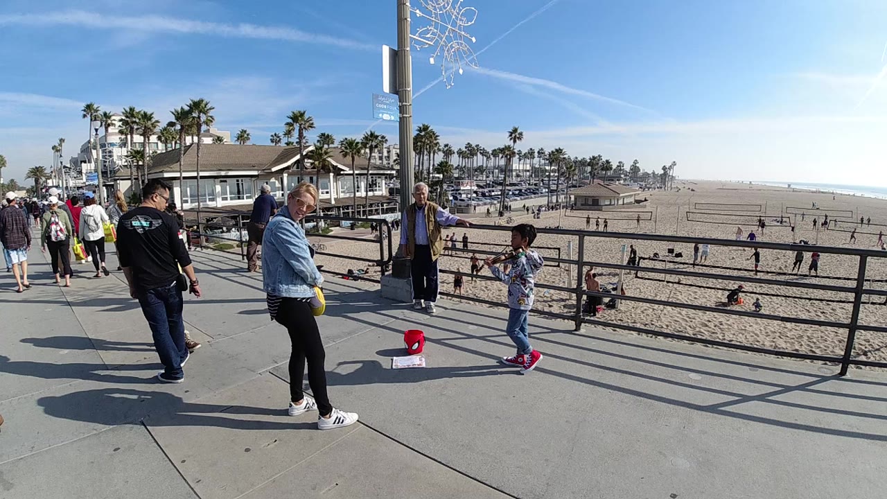 Adults and a child stand near a black railing on a Huntington Beach walkway, with sand, beachgoers, palm trees, and bright blue sky behind them.