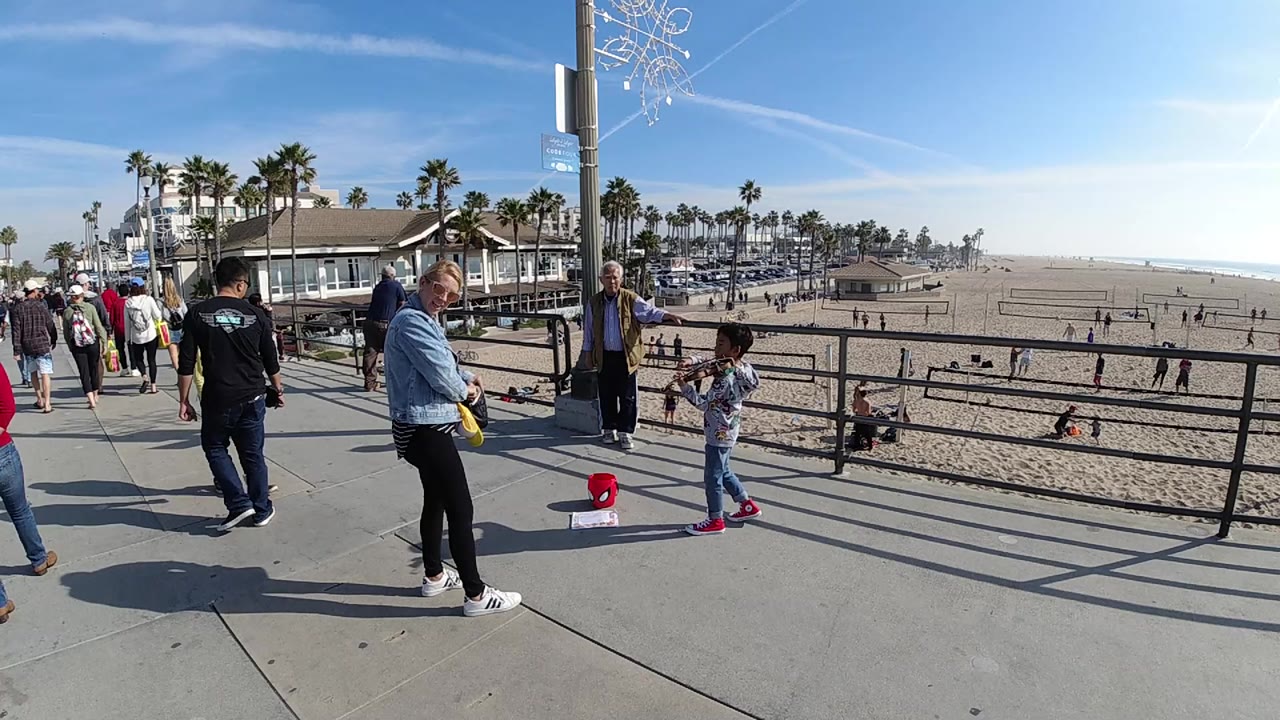 People walk and stand on a concrete path at Huntington Beach, California, with a black railing, sandy beach, palm trees, and bright sky beyond.