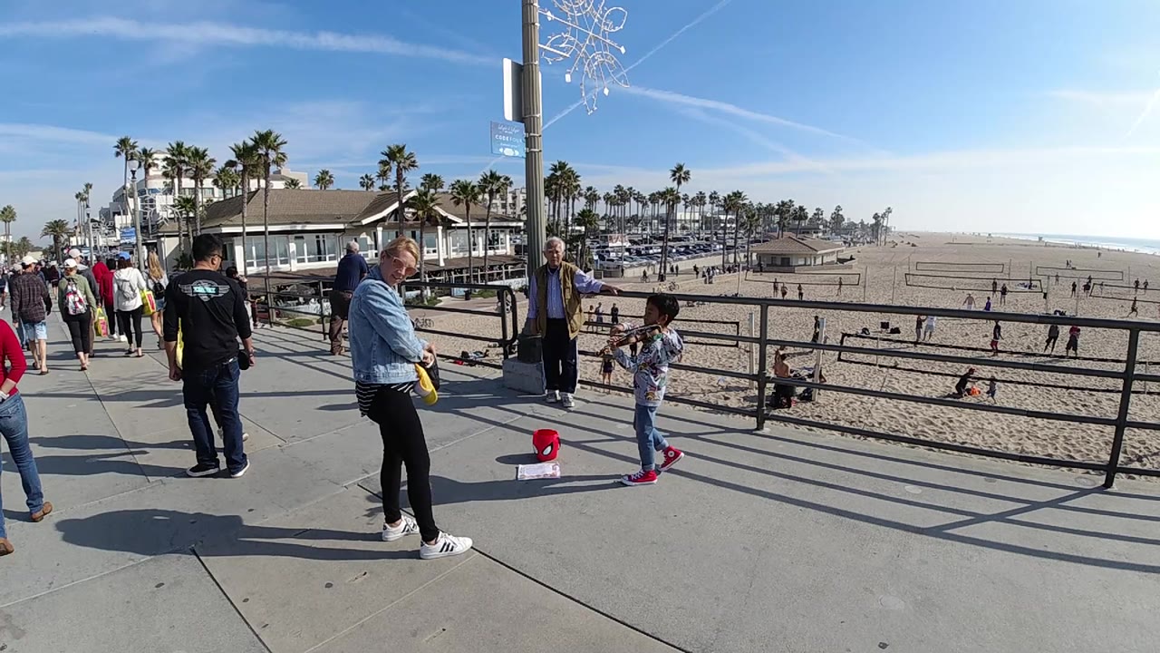 People occupy the foreground walkway at Huntington Beach, California, while the rail frames beachgoers, volleyball courts, palm trees, and a hazy ocean line.