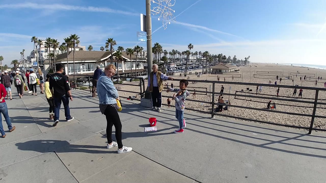 At 0:01 in Huntington Beach, the boy remains beside the railing with his violin as a woman in a denim jacket and other pedestrians face the performance against the beach backdrop.