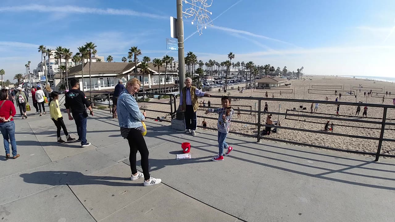 A young girl plays violin near a black railing on a Huntington Beach promenade as adults stand nearby and beachgoers cross the sand behind her.