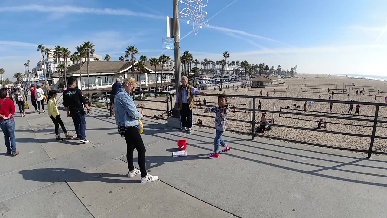 A wide Huntington Beach sidewalk view shows the young violinist beside her red bucket, with a man near a pole, people along the walkway, and beach activity beyond the railing.