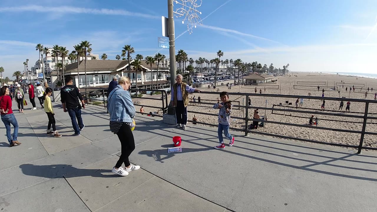 At 0:02, the young girl stands near the railing while the woman watches from the foreground, with Huntington Beach sand, scattered visitors, palms, and bright sky behind them.