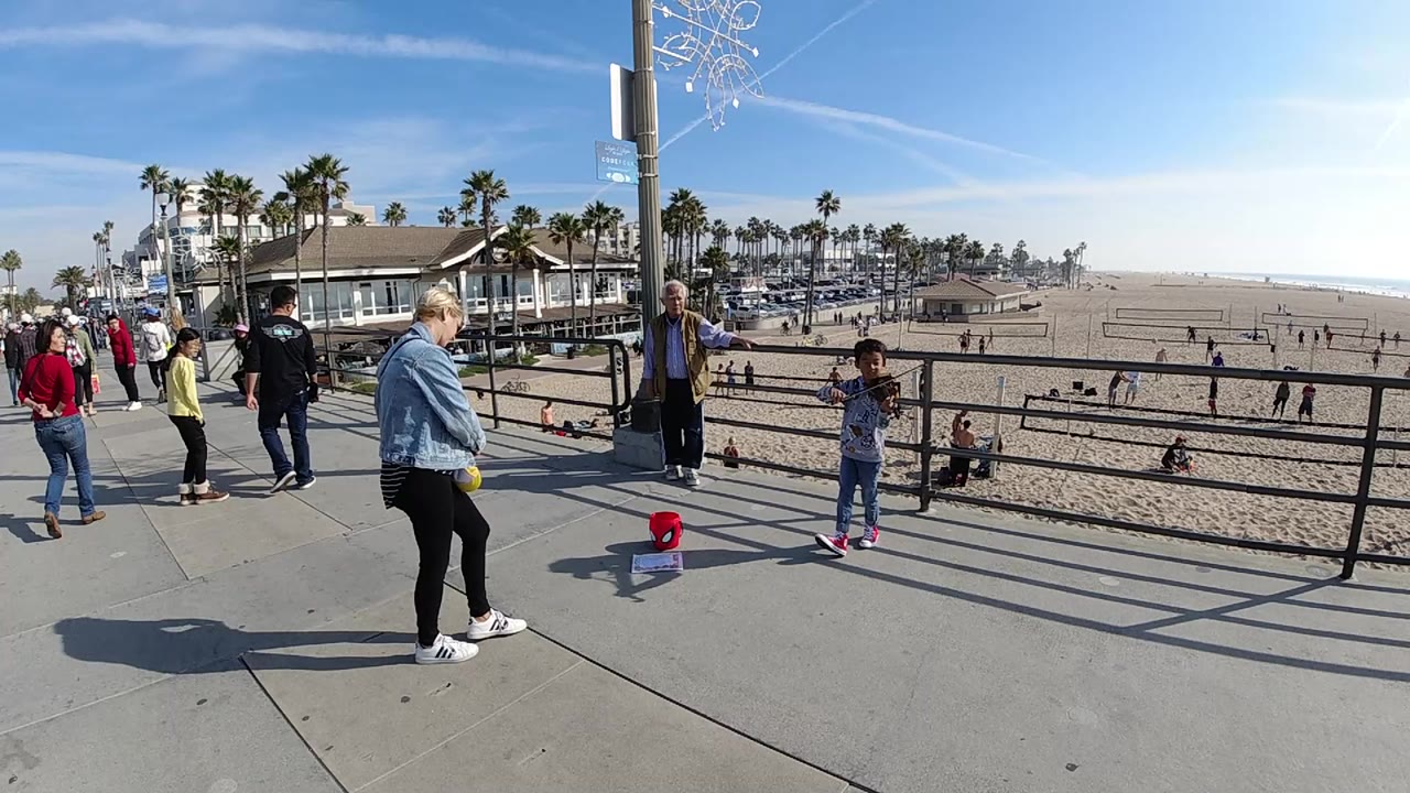 At Huntington Beach, a young boy stands near a red tip bucket by the promenade railing while adults watch and the sandy beach stretches behind him.