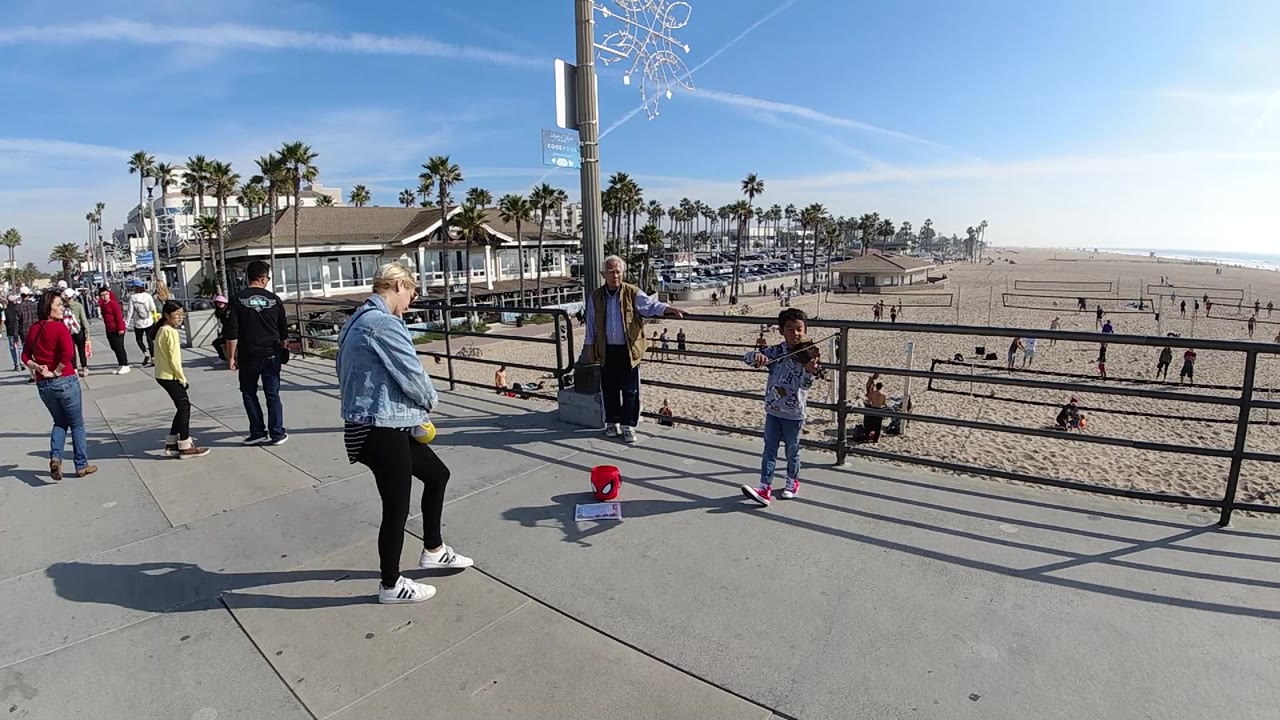 At Huntington Beach, the young performer faces the promenade area while several adults stand to the left and the wide sandy beach remains visible beyond the rail.