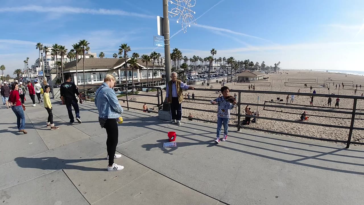At 0:00 in Huntington Beach, a child stands by a black beach railing while adults gather on the paved walkway and broad sand stretches behind them.