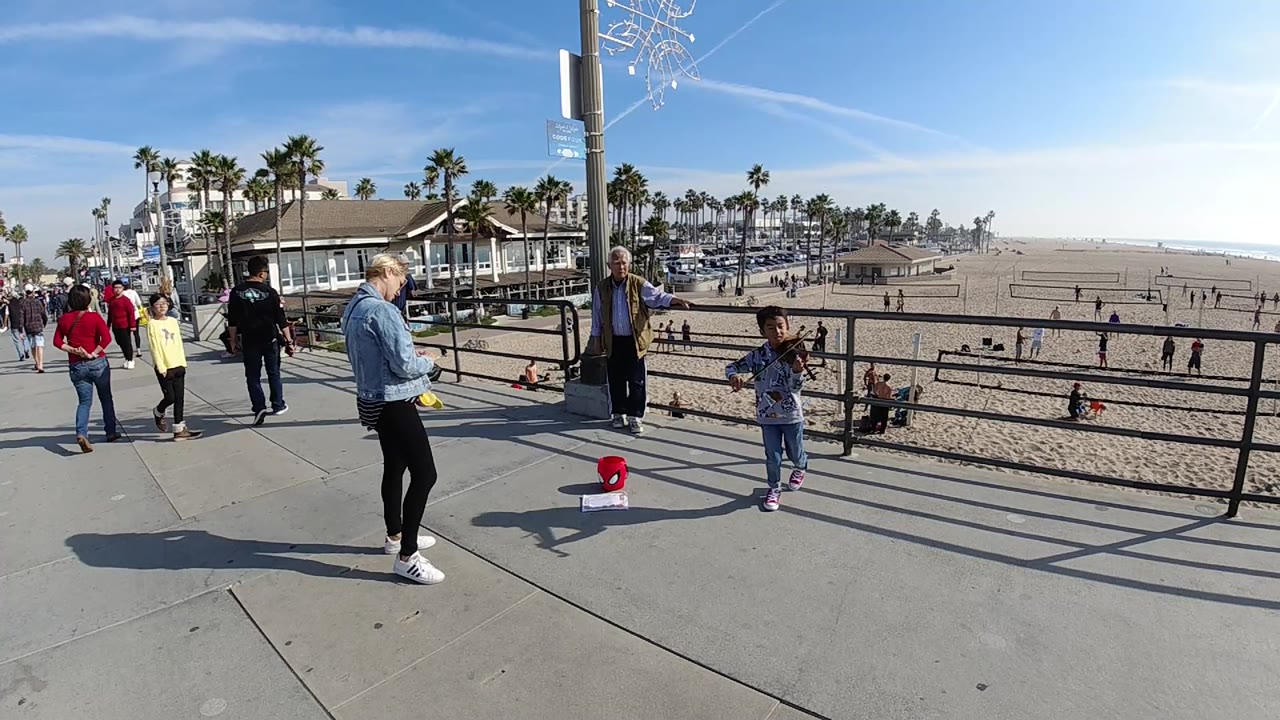 A very wide view at Huntington Beach shows the child violinist facing the walkway as pedestrians pass and palm trees line the distant beach area.