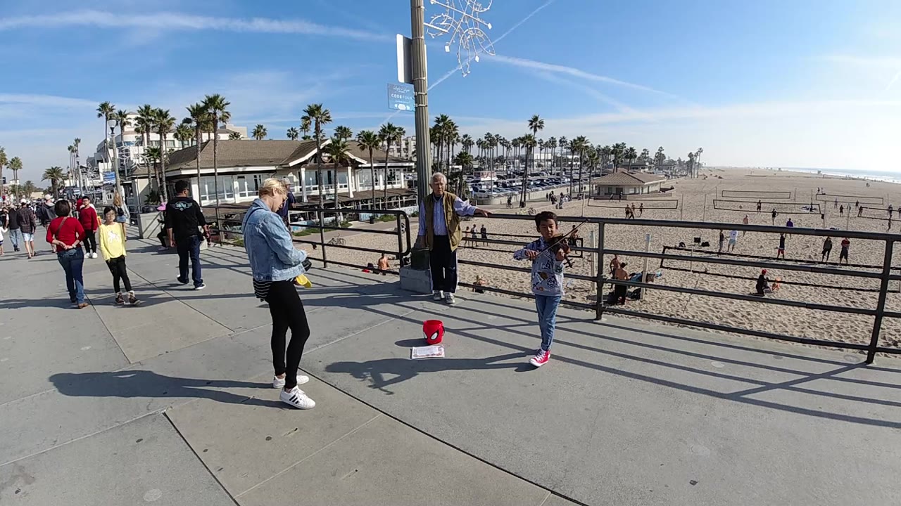 At Huntington Beach, California, a child stands near a railing as an adult in a denim jacket watches, with beach sand, pedestrians, palm trees, and blue sky behind them.