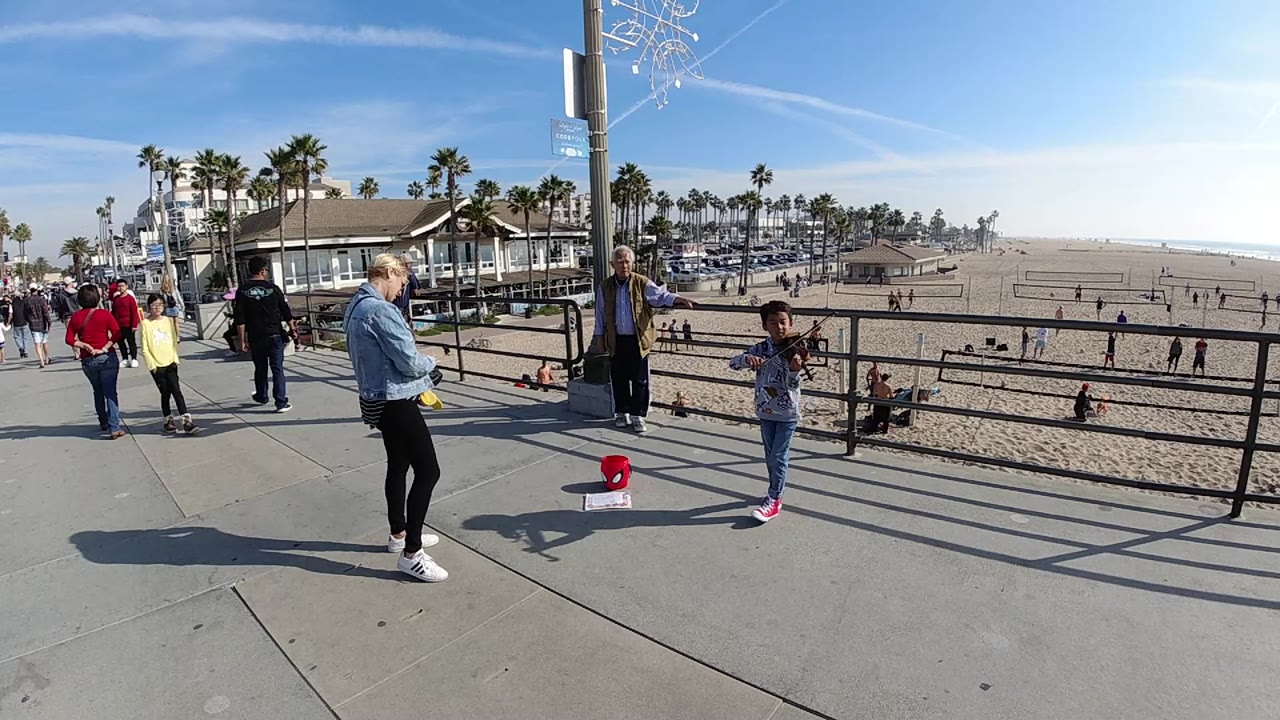 At 0:01 in Huntington Beach, California, the child remains near the railing with a red tip bucket on the pavement while beachgoers move along the walkway behind an adult observer.
