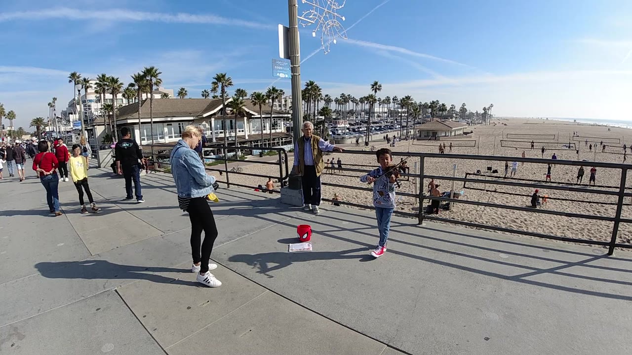 At Huntington Beach, California, the child performer stands to the right of the frame while the adult in denim and other pedestrians occupy the left side of the beach walkway.