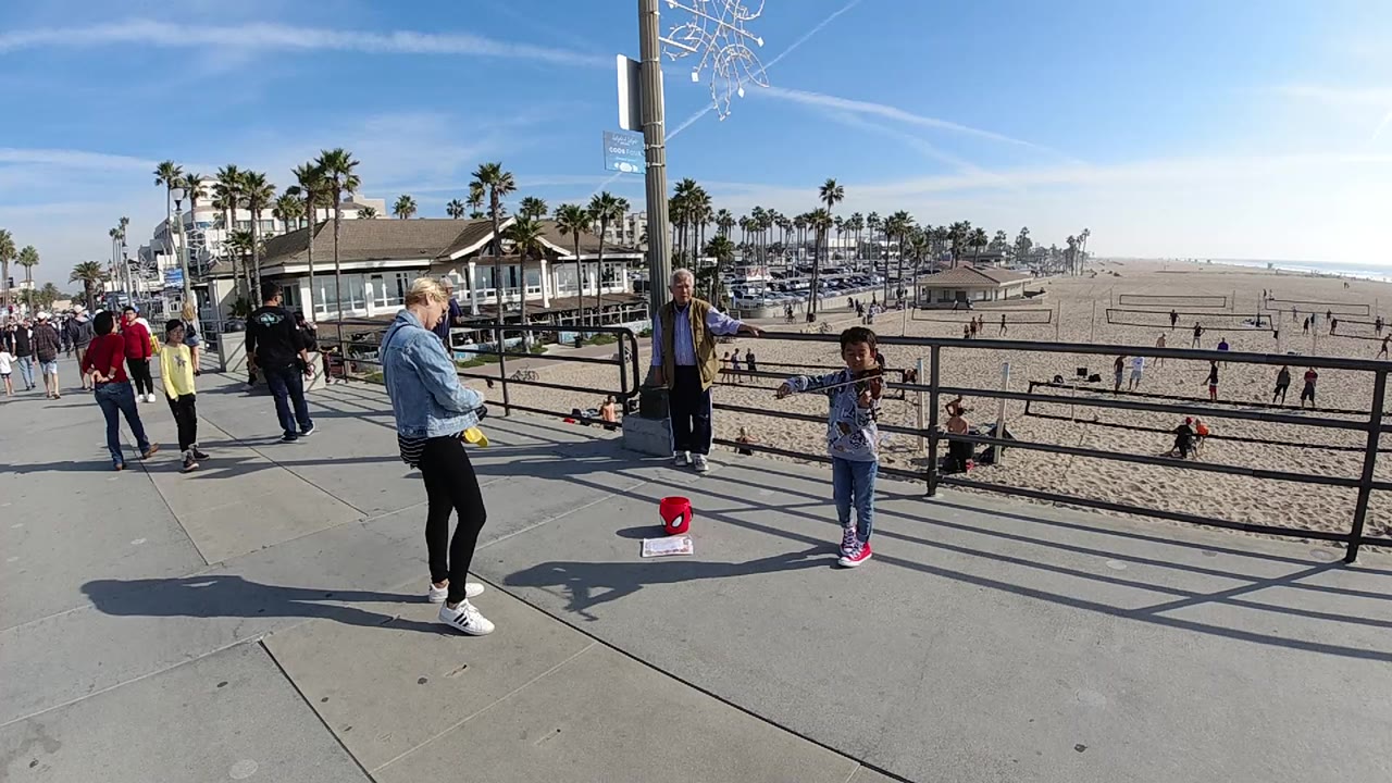 The Huntington Beach walkway scene continues with the boy near the railing, a woman standing opposite him, an older man behind them, and beach activity spread across the sand.