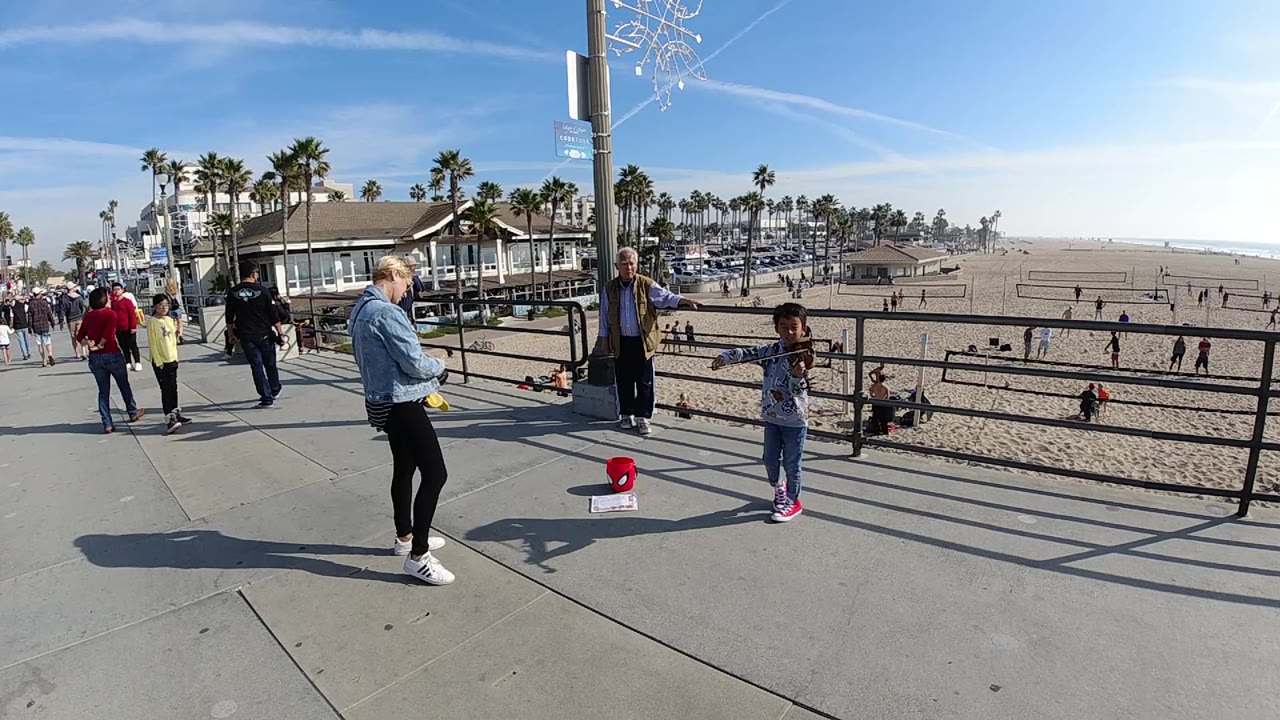 A wide Huntington Beach view frames the child performer near the right side, the woman and older man near the center, and a palm-lined beachfront with scattered people beyond the railing.