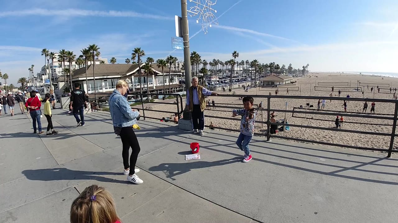 At Huntington Beach, California, a child plays violin beside a metal railing while an adult stands nearby, with a red tip bucket on the pavement and beach courts behind them.