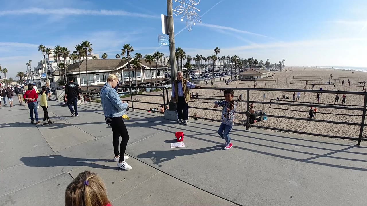The child violinist remains near the railing in Huntington Beach, California, with a woman facing the performer, an older adult by a pole, pedestrians to the left, and open sand in the background.
