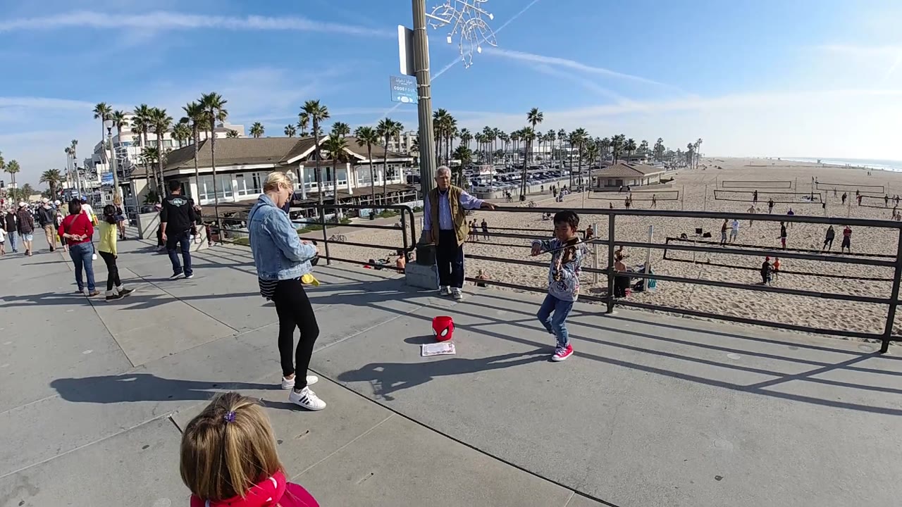 A wide view in Huntington Beach, California shows the child violinist at right, a woman in denim at center, an older adult near a pole, and beachgoers moving along the walkway and sand.