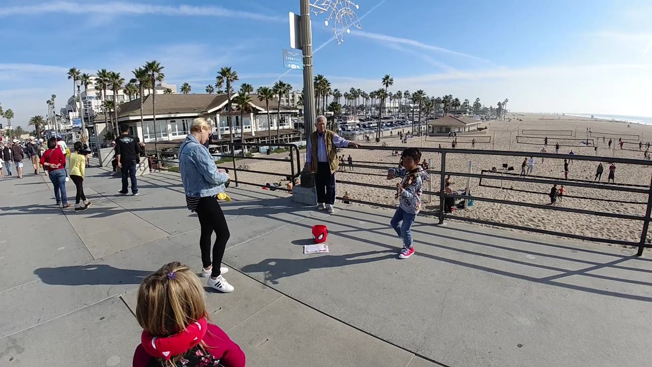 Child plays violin near a red tip bucket on a sunny beach walkway.