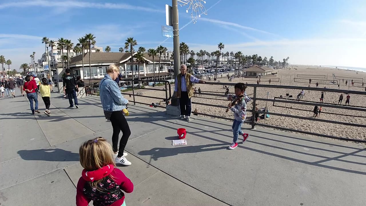 Wide frame of a Huntington Beach walkway with a child in the foreground, adults and another child near the railing, sandy beach, palms, and blue sky in the background.