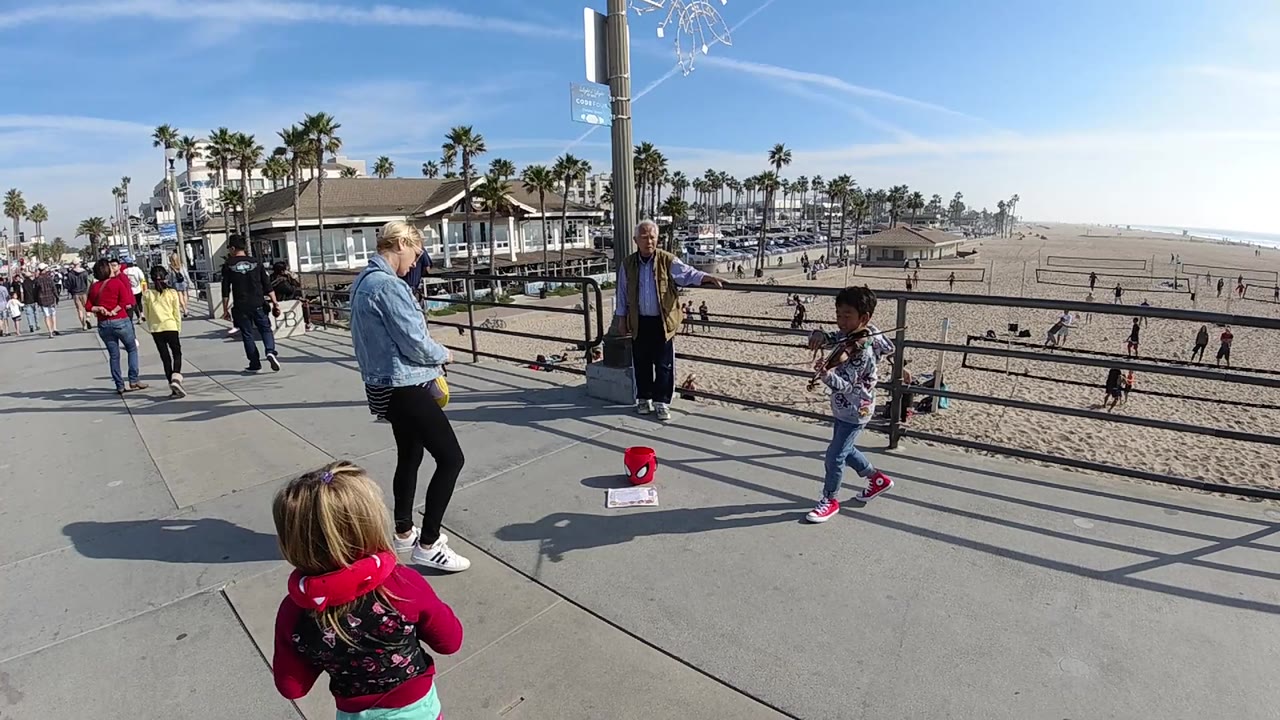 Wide Huntington Beach frame with a child moving near the railing, adults spaced across the walkway, beachgoers on the sand, palm trees, and a bright coastal sky.