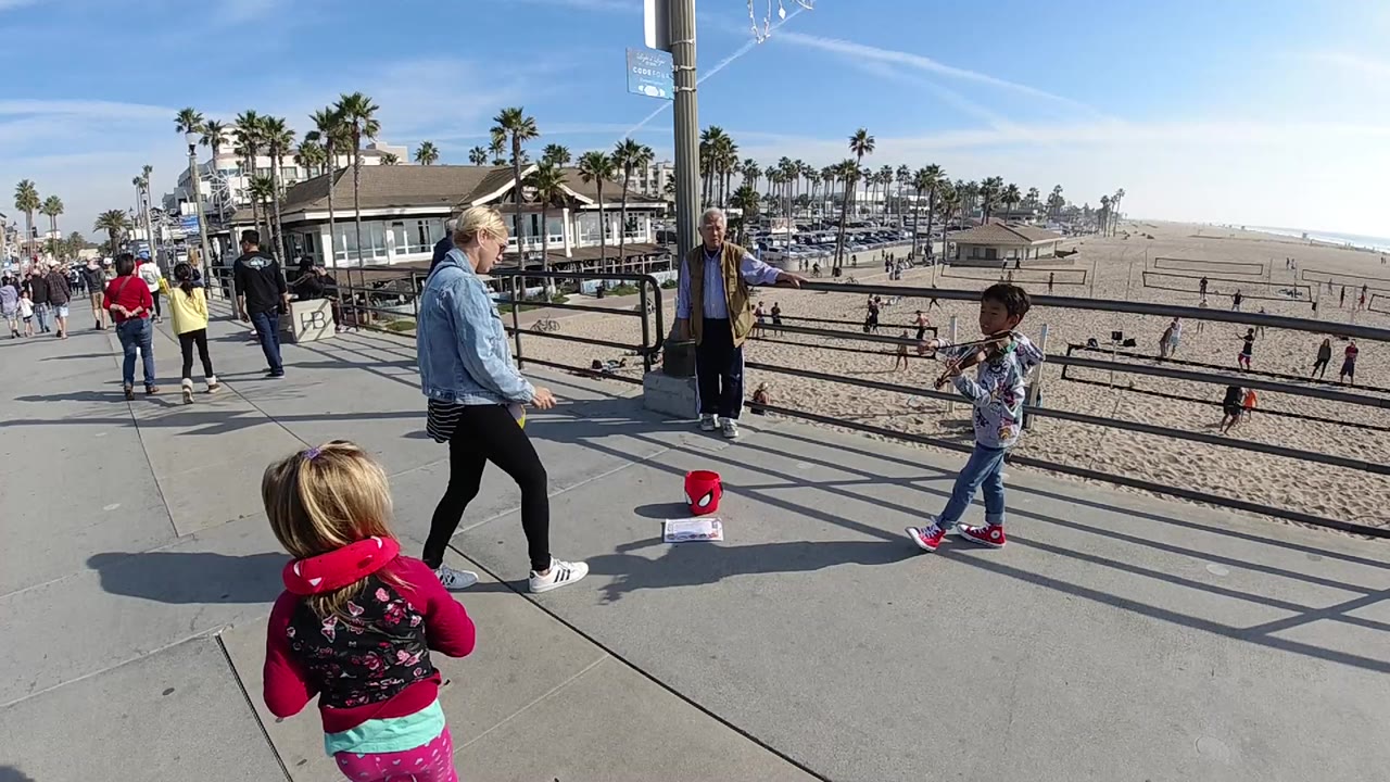 At 0:00 in Huntington Beach, adults and children stand and walk on a paved beachside walkway above sand, with palm trees, low buildings, railings, and bright blue sky in view.