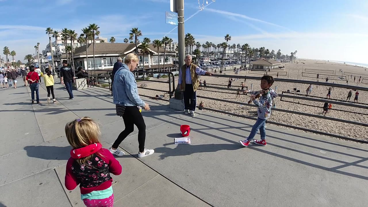 At 0:02 in Huntington Beach, the adult and child positions shift slightly on the walkway, with a red object on the pavement, rail shadows, palm trees, and open beach still visible.
