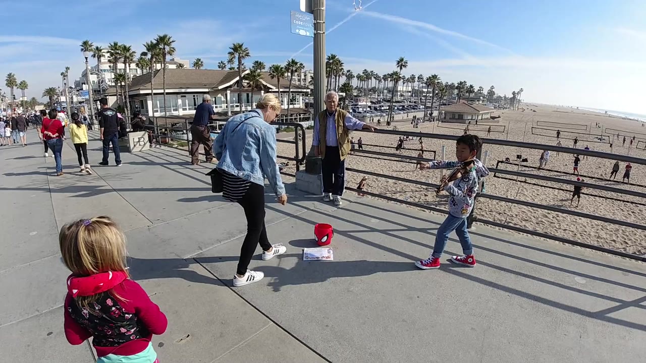 Huntington Beach overlook with an adult woman in a denim jacket moving toward the red tip bucket while the child violin player stands by the rail and beachgoers pass behind.