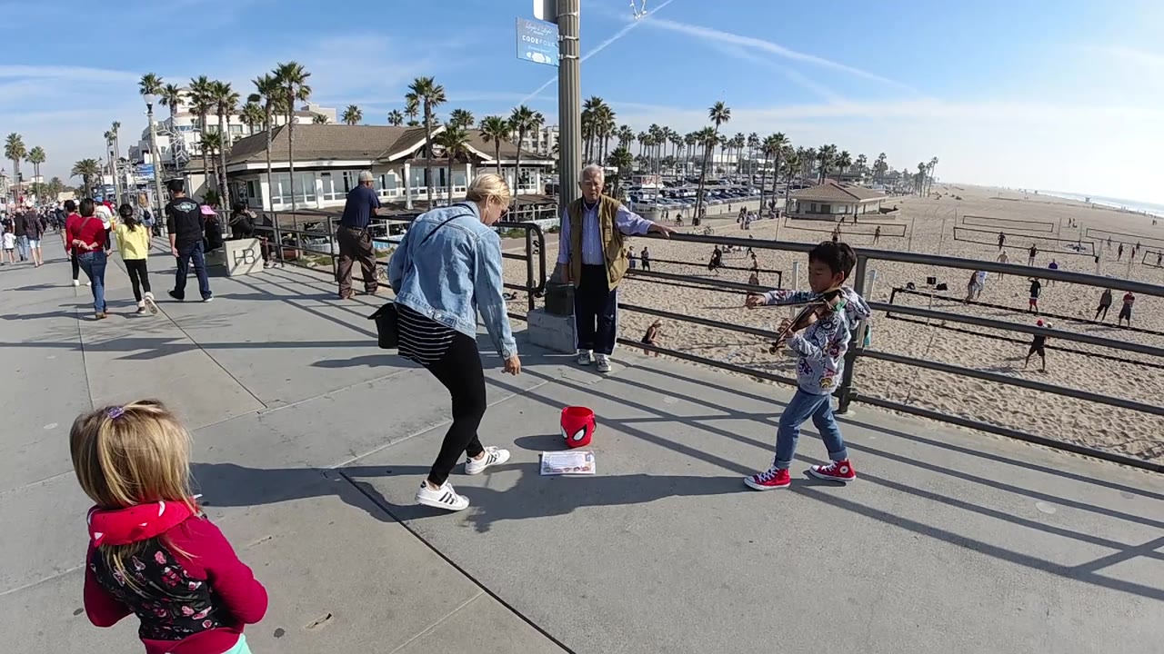 Huntington Beach promenade view with the child violin player at right, an adult woman leaning toward the tip bucket, other adults nearby, and the sandy beach visible through the railing.
