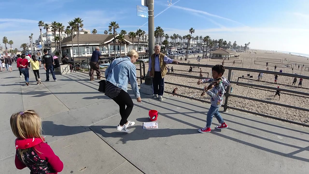 Woman bends toward a red tip bucket as a child plays violin beside the beach railing.