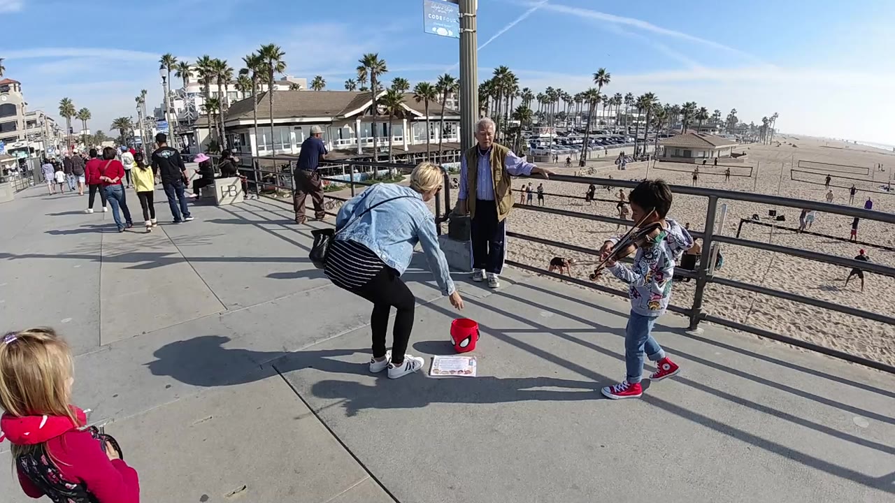 In Huntington Beach, a woman leans down toward a red donation bucket while a young violinist stands by the promenade railing with beach sand and palm trees behind them.