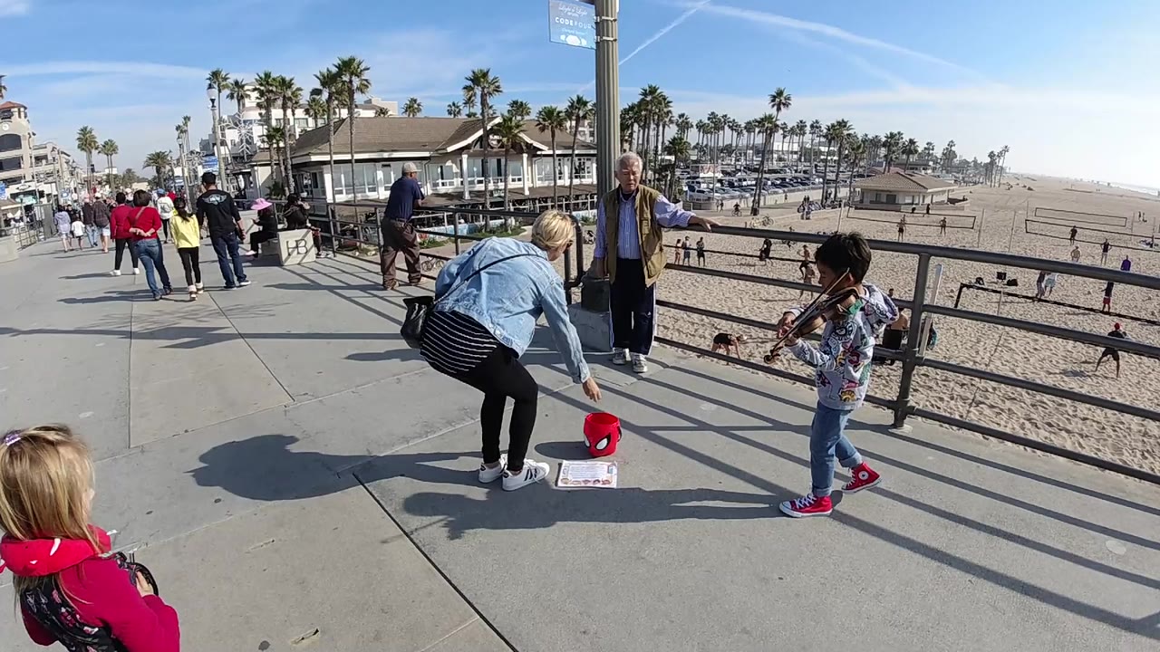 In Huntington Beach, the woman reaches lower toward the red bucket as the child violinist remains near the railing, with pedestrians moving along the walkway behind them.