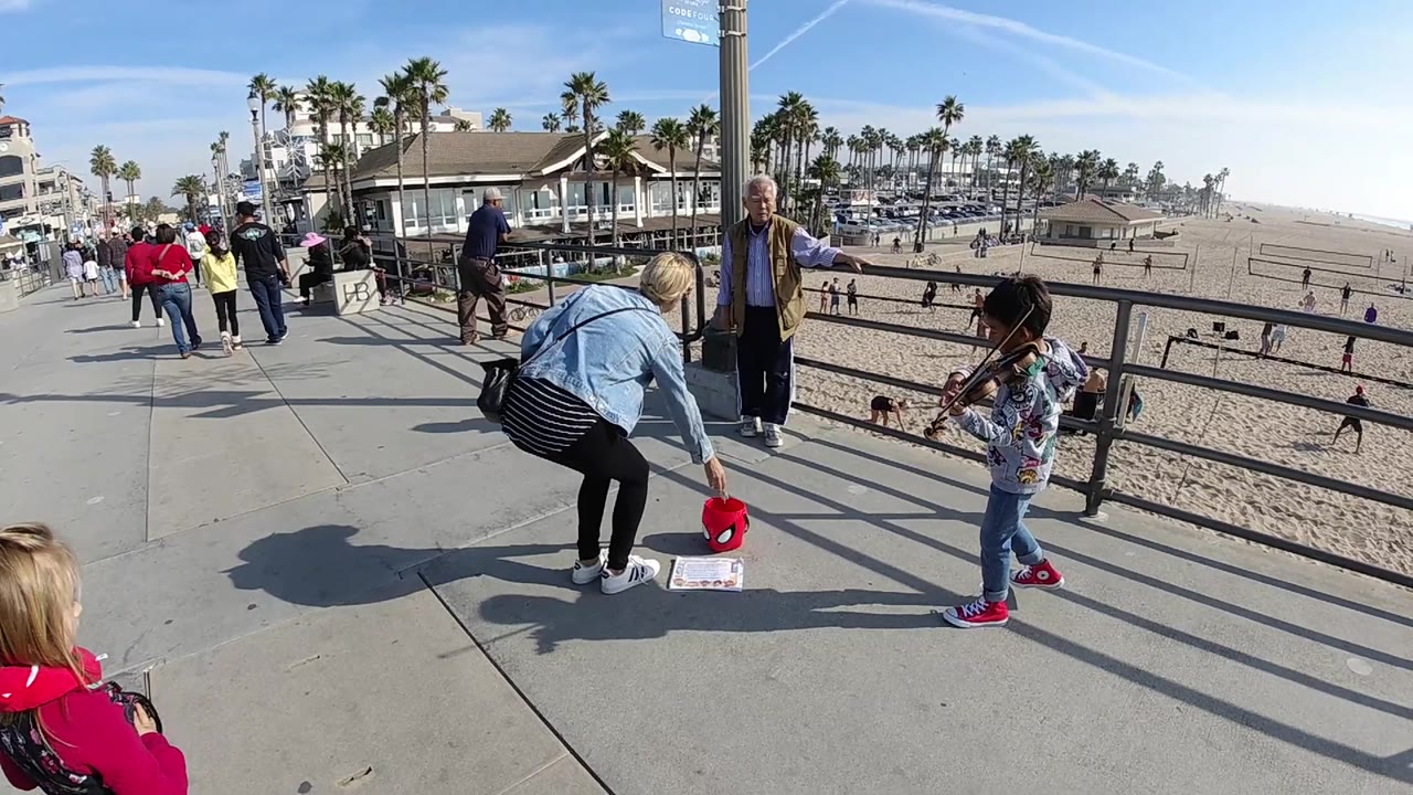 In Huntington Beach, the child violinist stands at the right of frame while the woman bends near the donation bucket, with beachgoers, railings, palms, and bright daylight in the background.