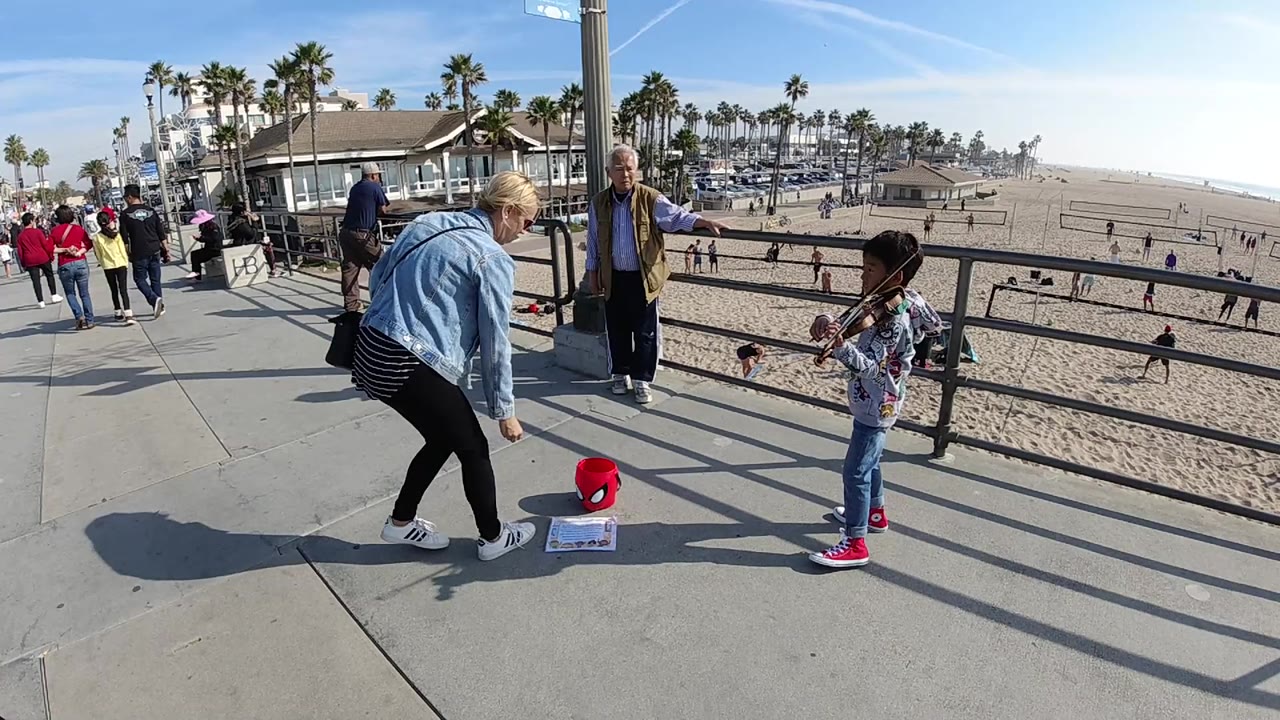Adult woman bends toward a red bucket as a child plays violin by the railing.