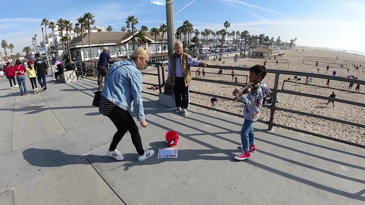 At Huntington Beach, California, the child violinist remains at the right of frame while the woman stays low near the bucket and the bright beach, rail shadows, and palm-lined background remain visible.